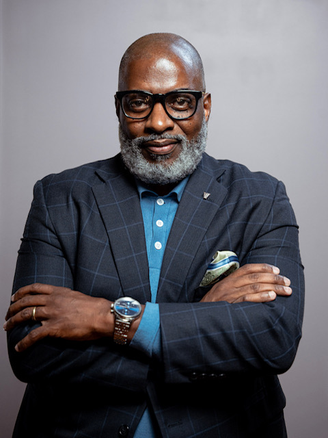 Avery Q. Muse, Sr., wearing glasses and a dark checked jacket, with his arms folded